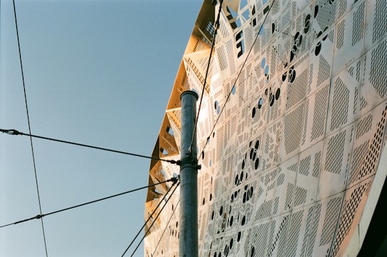 The image displays an intricate architectural facade with metallic panels featuring various perforated designs and patterns. A metallic pole intersects the view, with multiple cables connected to it, set against a clear blue sky.