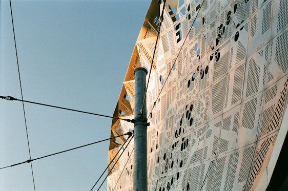 The image displays an intricate architectural facade with metallic panels featuring various perforated designs and patterns. A metallic pole intersects the view, with multiple cables connected to it, set against a clear blue sky.