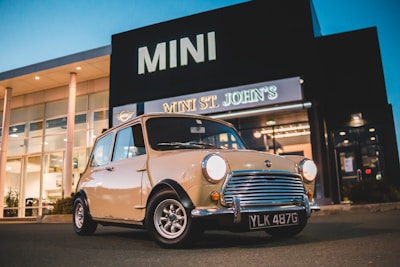 A vintage Mini Cooper car is parked in front of a building with signage reading 'MINI St. John's.' The car has a beige body with chrome accents and the headlights are on, illuminating the area slightly. The background shows a glass-fronted building under a clear evening sky.