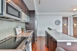 A modern kitchen features sleek dark cabinetry and stainless steel appliances. The countertop is a light, smooth surface, complemented by a neutral backsplash. A potted plant sits on the counter, and a wall clock is visible above the countertop. There is a wooden floor which adds warmth to the space.