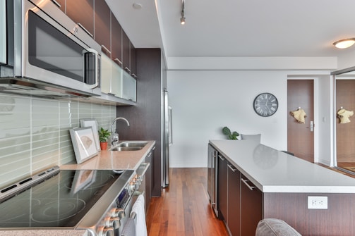 A modern kitchen features sleek dark cabinetry and stainless steel appliances. The countertop is a light, smooth surface, complemented by a neutral backsplash. A potted plant sits on the counter, and a wall clock is visible above the countertop. There is a wooden floor which adds warmth to the space.