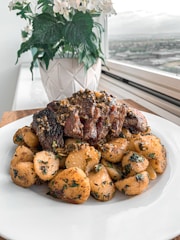 A plate filled with roasted potatoes and pieces of grilled meat, garnished with herbs. The dish is set next to a window with a view of a landscape. In the background, there is a decorative white pot with green leafy plants and white flowers.