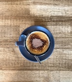 A cup of frothy cappuccino with cocoa powder sprinkled on top, placed on a saucer with a spoon, all set on a wooden table.