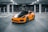 A sleek, sporty orange car with black accents is parked in an empty, dimly-lit parking garage. The vehicle has a modern design with aerodynamic curves and glossy finish, and is surrounded by concrete columns and reflective windows in the background.
