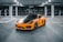 A sleek, sporty orange car with black accents is parked in an empty, dimly-lit parking garage. The vehicle has a modern design with aerodynamic curves and glossy finish, and is surrounded by concrete columns and reflective windows in the background.