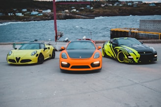 Three sports cars are parked by the waterfront on a concrete surface. The cars are vibrant yellow, orange, and black with neon green accents. The ocean and a rugged coastline with scattered houses are in the background.