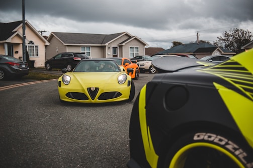 A dynamic scene features several sports cars in vibrant colors, including a prominent yellow car in the foreground, parked in a residential neighborhood. The sky appears overcast, hinting at possible rain.