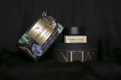 A Saint Jane Bright Repair eye cream container and its packaging are displayed. The packaging is adorned with floral patterns and features the product name in elegant gold lettering. Both the container and the box have a luxurious appearance, emphasizing premium skincare.