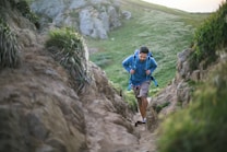 A person is hiking up a narrow, rocky trail surrounded by green vegetation. The individual is wearing a blue long-sleeve shirt and has a backpack, indicating they are prepared for an outdoor adventure. The surrounding landscape features grassy hills and jagged rocks, creating a natural and rugged setting.