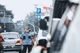 A man wearing a blue uniform and a cap stands on a busy city street. He appears to be managing or directing traffic, with several vehicles, including cars and motorcycles, visible around him. The man has a face mask lowered to his chin. There are multiple parking signs in the background, as well as trees and street lights, indicating an urban environment.