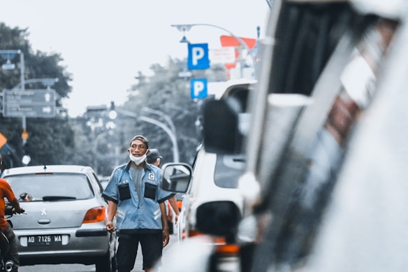 A man wearing a blue uniform and a cap stands on a busy city street. He appears to be managing or directing traffic, with several vehicles, including cars and motorcycles, visible around him. The man has a face mask lowered to his chin. There are multiple parking signs in the background, as well as trees and street lights, indicating an urban environment.