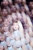 A kaleidoscope effect creates multiple overlapping images of a person with blond hair wearing a white, textured top. The image has a surreal, dreamlike quality with repeated faces and arms extending across the frame.