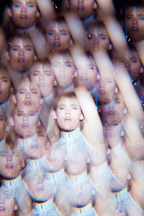 A kaleidoscope effect creates multiple overlapping images of a person with blond hair wearing a white, textured top. The image has a surreal, dreamlike quality with repeated faces and arms extending across the frame.