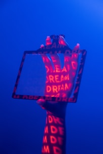 A hand is holding a transparent rectangular frame against a blue background. Red light projects the word 'DREAM' multiple times onto the hand and frame, creating a vivid contrast.