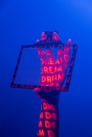 A hand is holding a transparent rectangular frame against a blue background. Red light projects the word 'DREAM' multiple times onto the hand and frame, creating a vivid contrast.