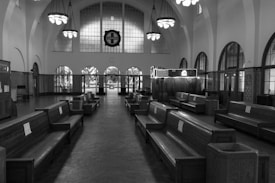 An empty, historical-looking train station interior features long wooden benches aligned in rows. Arched windows and high ceilings with hanging chandeliers contribute to the vintage atmosphere.