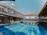 A luxurious swimming pool with clear blue water is surrounded by ornate architecture. The building features intricate railings and several classical statues. There are outdoor seating areas with tables and chairs under umbrellas.