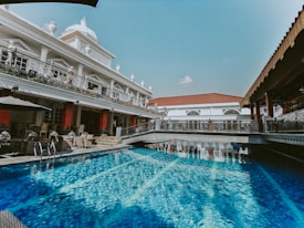 A luxurious swimming pool with clear blue water is surrounded by ornate architecture. The building features intricate railings and several classical statues. There are outdoor seating areas with tables and chairs under umbrellas.