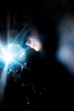 A person is partially obscured by shadows, holding a reflective disco ball emitting bright lights. The scene is dimly lit, creating a mysterious and intriguing atmosphere.