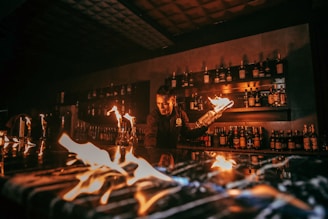 A bartender is performing a fire show behind a bar counter. Several bottles of liquor are visible on shelves in the background. The flames create an intense and captivating atmosphere.