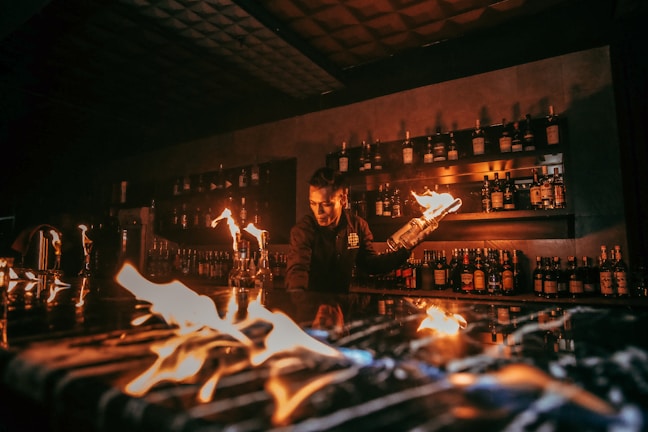 A bartender is performing a fire show behind a bar counter. Several bottles of liquor are visible on shelves in the background. The flames create an intense and captivating atmosphere.