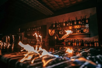A bartender is performing a fire show behind a bar counter. Several bottles of liquor are visible on shelves in the background. The flames create an intense and captivating atmosphere.