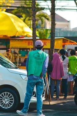 A group of people stands near a street food stall with a prominent yellow canopy. In the foreground, a man wearing a green vest and a baseball cap faces away, possibly monitoring the area. A white car is parked on the street beside them. The scene suggests a busy urban setting with trees and buildings in the background.