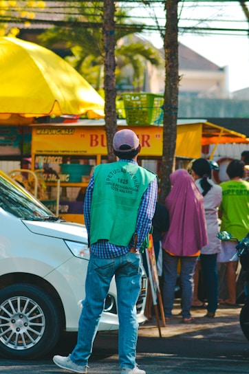 A group of people stands near a street food stall with a prominent yellow canopy. In the foreground, a man wearing a green vest and a baseball cap faces away, possibly monitoring the area. A white car is parked on the street beside them. The scene suggests a busy urban setting with trees and buildings in the background.