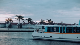 A serene waterfront scene with a boat on the water and palm trees lining the shore. In the background, there is a large hotel complex with distinct red roofs and white walls. The sky is overcast, hinting at an approaching sunset.