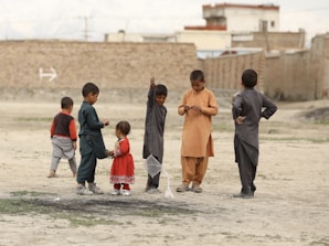 A group of children is playing outdoors on a dirt field. Some are wearing traditional clothing in various colors. A young girl in a red dress stands among the boys. The children are engaged in different activities, with one holding a small net. In the background, there are brick walls and some distant buildings.