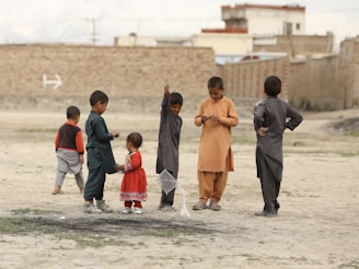 A group of children is playing outdoors on a dirt field. Some are wearing traditional clothing in various colors. A young girl in a red dress stands among the boys. The children are engaged in different activities, with one holding a small net. In the background, there are brick walls and some distant buildings.