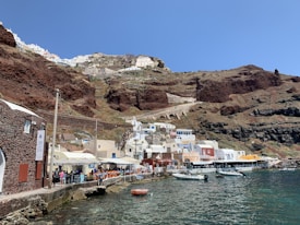 The image depicts a scenic coastal town built into rugged, reddish-brown cliffs. The town features whitewashed buildings with flat roofs and some colorful facades, including shades of beige, yellow, and red. A line of waterfront cafes with outdoor seating runs along a pier, and several small boats are moored in the clear, blue-green water. The sky is clear and blue, adding to the serene atmosphere.