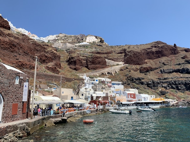 The image depicts a scenic coastal town built into rugged, reddish-brown cliffs. The town features whitewashed buildings with flat roofs and some colorful facades, including shades of beige, yellow, and red. A line of waterfront cafes with outdoor seating runs along a pier, and several small boats are moored in the clear, blue-green water. The sky is clear and blue, adding to the serene atmosphere.