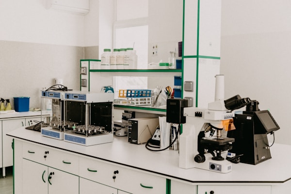 A laboratory workspace featuring a variety of equipment including a microscope, other analytical devices, and shelves with bottles and laboratory supplies. The room is organized and well-lit with clean surfaces and cabinetry.