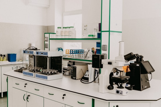 A laboratory workspace featuring a variety of equipment including a microscope, other analytical devices, and shelves with bottles and laboratory supplies. The room is organized and well-lit with clean surfaces and cabinetry.