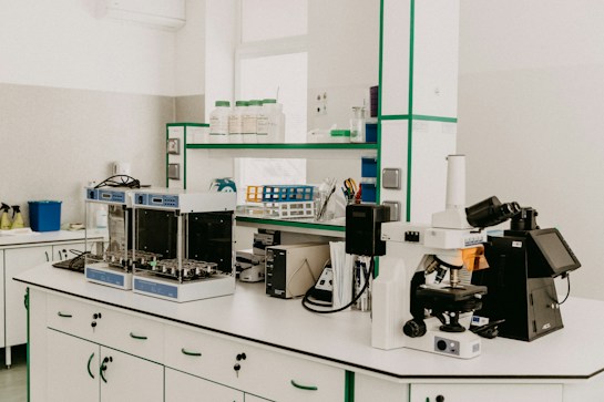 A laboratory workspace featuring a variety of equipment including a microscope, other analytical devices, and shelves with bottles and laboratory supplies. The room is organized and well-lit with clean surfaces and cabinetry.