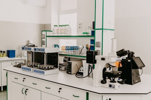 A laboratory workspace featuring a variety of equipment including a microscope, other analytical devices, and shelves with bottles and laboratory supplies. The room is organized and well-lit with clean surfaces and cabinetry.