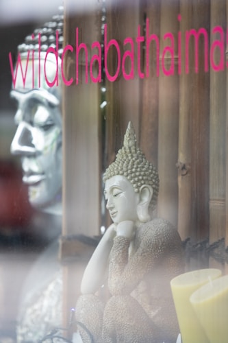 A serene sculpture of a meditative figure with curly hair is placed beside an array of decorative items, including wooden elements and a pair of yellow candles. The scene is viewed through a glass window with the words 'wildchabathaimassage' displayed in pink lettering. The soft lighting and reflections add a tranquil atmosphere.