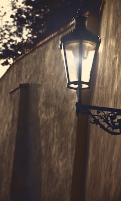 A vintage street lamp is mounted on an old textured wall with intricate wrought-iron brackets. The light casts a soft glow in a dimly lit setting, with shadows and trees in the background.
