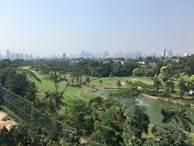 A lush, green golf course with well-maintained fairways and several water features surrounded by dense trees. In the background, a cityscape with numerous tall buildings is visible under a clear sky.