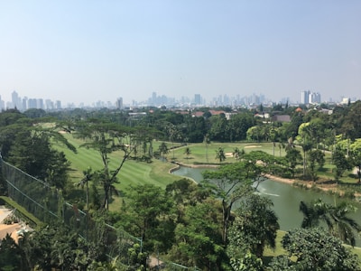 A lush, green golf course with well-maintained fairways and several water features surrounded by dense trees. In the background, a cityscape with numerous tall buildings is visible under a clear sky.