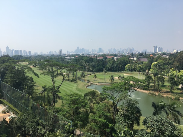 A lush, green golf course with well-maintained fairways and several water features surrounded by dense trees. In the background, a cityscape with numerous tall buildings is visible under a clear sky.