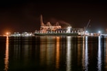 A large cargo ship is docked at a bustling port during nighttime, with cranes towering over it. The scene is illuminated by artificial lights, casting reflections on the calm water. Containers are stacked high on the ship, indicating active maritime trade.
