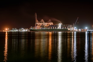 A large cargo ship is docked at a bustling port during nighttime, with cranes towering over it. The scene is illuminated by artificial lights, casting reflections on the calm water. Containers are stacked high on the ship, indicating active maritime trade.
