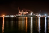 A large cargo ship is docked at a bustling port during nighttime, with cranes towering over it. The scene is illuminated by artificial lights, casting reflections on the calm water. Containers are stacked high on the ship, indicating active maritime trade.