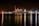 A large cargo ship is docked at a bustling port during nighttime, with cranes towering over it. The scene is illuminated by artificial lights, casting reflections on the calm water. Containers are stacked high on the ship, indicating active maritime trade.
