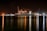 A large cargo ship is docked at a bustling port during nighttime, with cranes towering over it. The scene is illuminated by artificial lights, casting reflections on the calm water. Containers are stacked high on the ship, indicating active maritime trade.