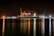 A large cargo ship is docked at a bustling port during nighttime, with cranes towering over it. The scene is illuminated by artificial lights, casting reflections on the calm water. Containers are stacked high on the ship, indicating active maritime trade.