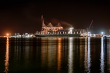 A large cargo ship is docked at a bustling port during nighttime, with cranes towering over it. The scene is illuminated by artificial lights, casting reflections on the calm water. Containers are stacked high on the ship, indicating active maritime trade.