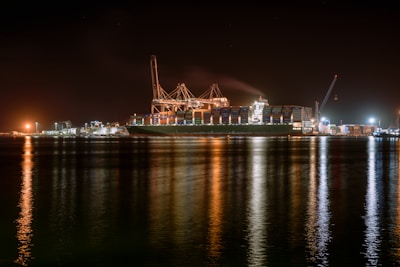 A large cargo ship is docked at a bustling port during nighttime, with cranes towering over it. The scene is illuminated by artificial lights, casting reflections on the calm water. Containers are stacked high on the ship, indicating active maritime trade.